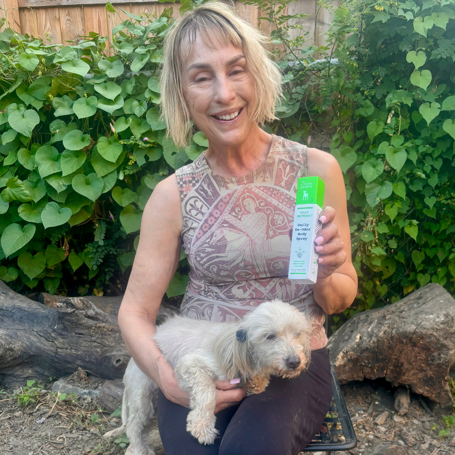 Woman holding a small dog and a product package outdoors with greenery in the background